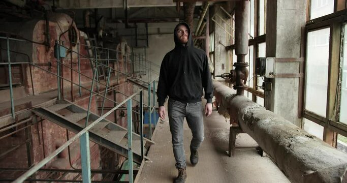 a young bearded man in a hood goes to the destroyed factory. around shabby walls, torn wires, rusty pipes and railing.