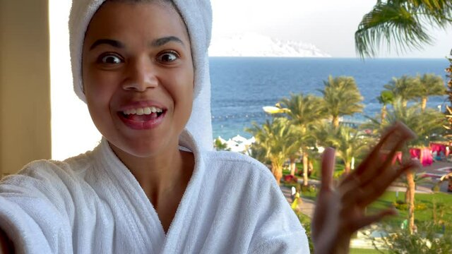 Celebrate life. Excited young woman in bathrobe having video conference call with friends while standing on the hotel balcony with sea view. Vacation, technology concept
