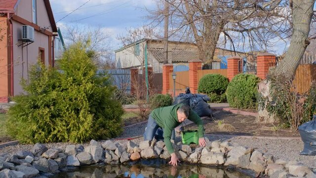 Mature adult caucasian man cleans a garden pond from water plants and falling leaves and puts it in a trash bag. Spring seasonal pond care after winter. 
