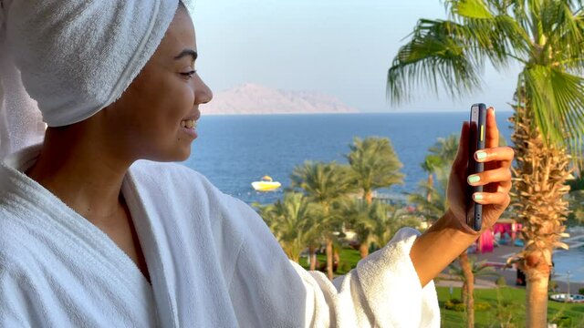 Bye bye. Happy young woman in bathrobe having video conference call with friends, saying goodbye and blowing a kiss while standing on the hotel balcony with sea view. Vacation, technology concept