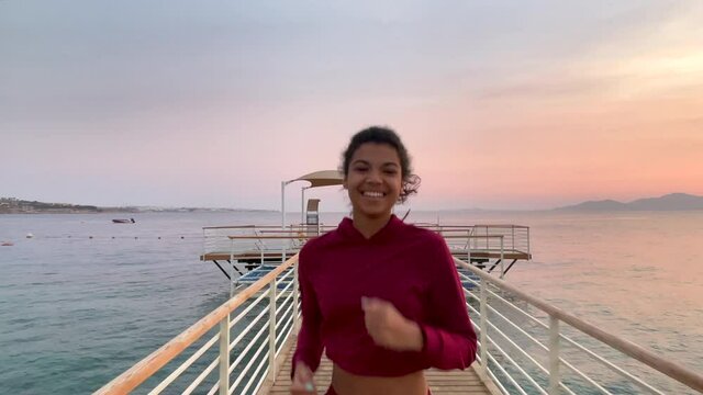 Early morning. Front view of happy young woman smiling while quickly running along the pier at sunrise. Slow motion. Healthy lifestyle, nature concept
