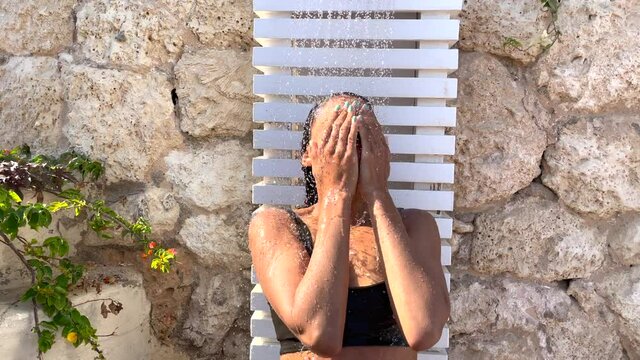 Stay fresh. Young woman in swimsuit enjoys taking outdoor shower, opens her eyes and laughing. Camera close up. Skincare on vacation, relax, summertime concept