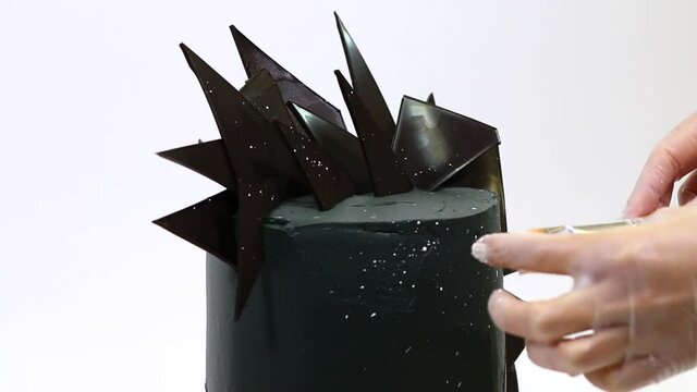 Professional pastry chef decorates stylish brutal black cake with pieces of chocolate. Abstract cake in the style of space on white background. 
