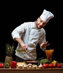 Handsome chef in white uniform pours butter on chopped vegetables in a frying pan