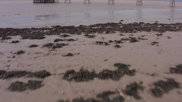 Aerial tilting up from muddy beach past clumps of seaweed to reveal Victorian Clevedon Pier on the Severn Estuary on a sunny day