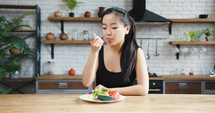 Asian woman sitting at table, looking sad and bored with diet not wanting to eat salad. Modern kitchen interior