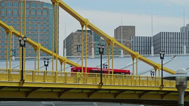 A long shot view of a red Pittsburgh city bus traveling on the yellow Rachel Carson Bridge. The downtown skyline seen in the distance.  	
