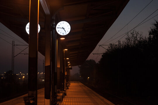 Analog clock on the platform of a commuter train station at night, during a dark sunset evening, indicating the precise time, in front of a deserted track with nobody and no train. - Powered by Adobe