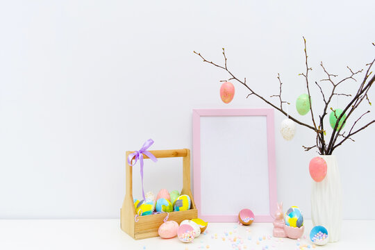 Trendy colorful Easter composition with hand painted eggs in yellow and grey colors in basket, geometric vase, confetti, twigs, pink Easter bunny. Blank photo frame, mockup with copy space for text.
