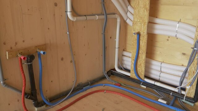 CLOSE UP: Recuperation system and other tubing runs across a room inside a wooden house under construction. Floor heating fuse box stands in the unfinished utility room full of plastic conduits.