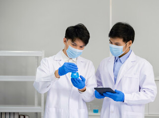 Two Scientists discuss about blue liquid chemicals in Erlenmeyer flask. Asian doctor use tablet computer to research chemical compound on the internet. Covid-19 vaccine testing in laboratory.