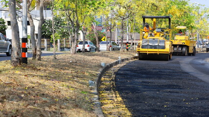 Obraz premium Yellow road roller on the road. On a paved road that is being repaired or rebuilt, two yellow road rollers are working. On a green wood background with yellow flowers with copy space. Selective focus
