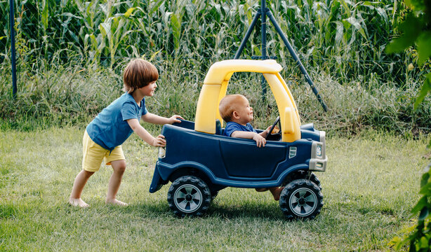 Brother helps brother drive the children's car. The boys are playing in the garden with a toy car. Brotherly help and having fun.