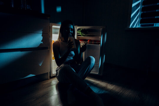 A Hungry Girl Sits With Her Elbows On The Open Refrigerator And Drinks Milk. The Image Was Shot In A Low Key To Convey Night Time.