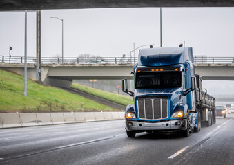 Blue classic industrial big rig semi truck transporting covered cargo on flat bed semi trailer running on the wide highway road at wet rain weather