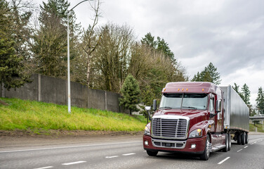 Fototapeta na wymiar Burgundy low cab big rig semi truck transporting cargo in low profile covered semi trailer running on the multiline highway road with hill and fence on the side