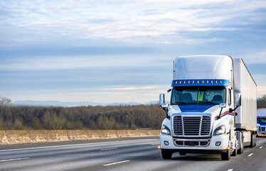 Day cab big rig white semi truck with roof spoiler transporting cargo in dry van semi trailer running in front of truck convoy on the wide multiline highway road