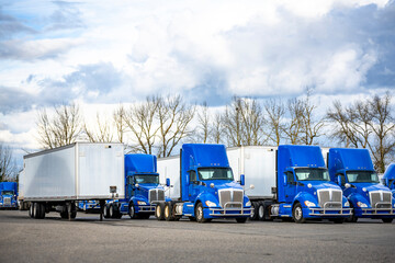 Day cab blue big rig semi trucks tractors and dry van semi trailers standing at row on industrial parking lot waiting for next freight route