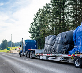 Huge blue classic big rig semi truck transporting covered heavy commercial cargo on step down semi...