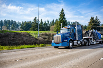 Powerful blue big rig semi truck tractor transporting heavy covered commercial cargo on step down...