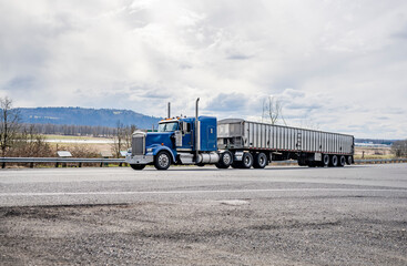 Powerful classic blue big rig American semi truck transporting cargo in low profile long bulk semi...