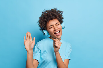 Indoor shot of carefree joyful African American woman keeps hand near mouth as if microphone sings song along closes eyes from pleasure listens music via wireless headphones isolated over blue wall
