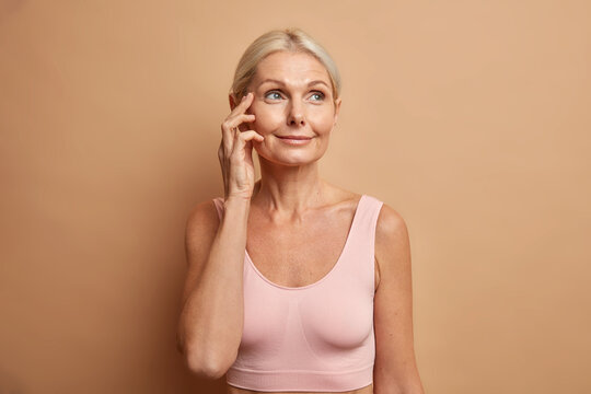 Photo of charming middle aged woman touches face has healthy skin after cosmetic procedures or facial treatments wears cropped top poses with bare shoulders isolated over brown studio background.