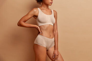 Sideways shot of slim woman wears cropped top and panties demonstrates perfect figure after weigh loss poses against beige studio background - obrazy, fototapety, plakaty