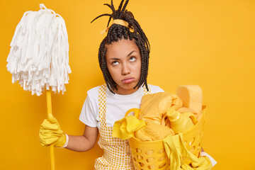 Housework concept. Unhappy housewife feels tired holds mop laundry basket does housework dressed in...