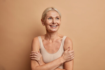 Attractive positive wrinkled fifty years old woman looks gladfully above keeps arms foded has well...