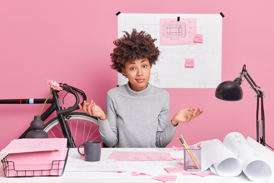 Hesitant talented female architect spreads palms poses confused at desktop doesnt know how to improve design sketches isolated over pink background tries to decice how improve productivity of work