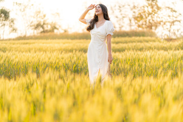 Obraz premium Happy young beautiful Asian woman in white dress standing in the barley field enjoying her time outdoor alone during sunset