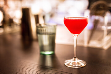 A bright red alcoholic cocktail in a coupe glass and a glass of water at the bar counter. A horizontal lifestyle photo with shallow depth of field and copy space.