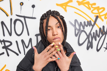 Self confindent cool teenager looks seriously at camera raises hands bites lips has dreadlocks hairstyle wears fashionable clothes poses against graffiti wall. Youth fashion teenage lifestyle concept - obrazy, fototapety, plakaty