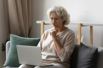 Serious older woman using laptop for video call from home, making shh gesture at webcam. Focused...