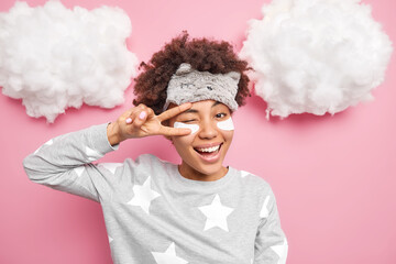 Positive African American woman smiles gladfully makes peace gesture over eye winks eyes applies collagen patches under eyes dressed in pajama isolated over pink background white clouds above