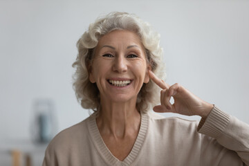 Portrait of happy senior woman pointing finger at her ear, looking at camera and smiling. Excited...