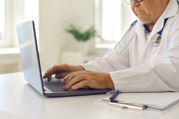 Senior doctor using laptop, giving online consultations to patients or entering data into system