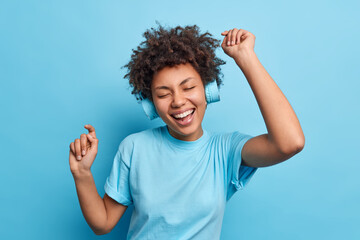 Joyful relaxed African American girl enjoys favorite playlist listens music via wreless headphones raises arms dressed casually isolated over blue background. People hobby and lifestyle concept