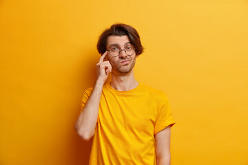 Photo of thoughful young European keeps finger on temple imagines something wears round spectacles and casual t shirt isolated over yellow studio background makes important decision picks variant