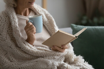 Old retired 70s lady reading book at home, drinking hot tea, resting on sofa in cozy living room....
