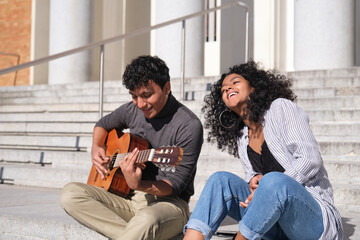 A young latin couple playing the guitar and singing sitting on stairs. Millennial generation.