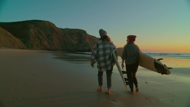 Two women walk with surfboards on empty epic beach towards surfing spot. Sunrise or sunset amazing views. Camera follows casual female surfers. Two friends have fun at beach