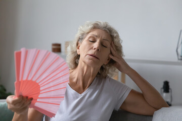 Mature senior woman exhausted with heat, swelter, summer weather, waving hand fan at home, cooling in too hot air. Tired unhappy elderly 60s lady suffering from hormone imbalance menopause symptoms