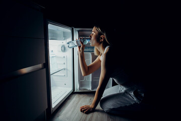 A young European woman suffers from the summer heat and lack of air conditioning at home. The girl in sweat drinks fresh water from the refrigerator.