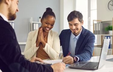 Happy african american woman watches as her caucasian husband signs a contract to rent or buy property in the office. Interracial clients at a meeting with a realtor or bank manager.