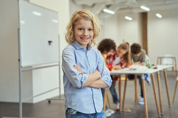 Smart guy. Portrait of joyful boy smiling at camera, standing with arms crossed while posing for camera during crafting classes. Kids working on diy robot in the background