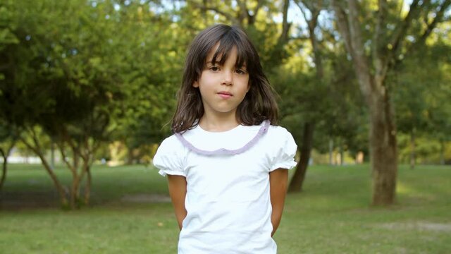 Pretty cute little black haired girl standing in city park, looking at camera and smiling. Kid enjoying leisure time outdoors in summer. Medium shot. Childhood concept