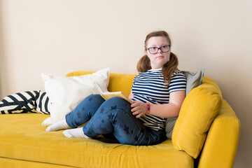 Cute teenager girl with glasses with Down syndrome sitting on sofa at home and reading book. Domestic life of people with disabilities. Selective focus.