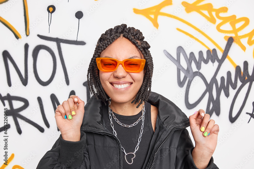 Wall mural Photo of joyful teenage girl bites lips dances carefree dressed in fashionable clothes expresses positive emotions poses against graffiti wall foolishes around. Youth lifestyle modern generations